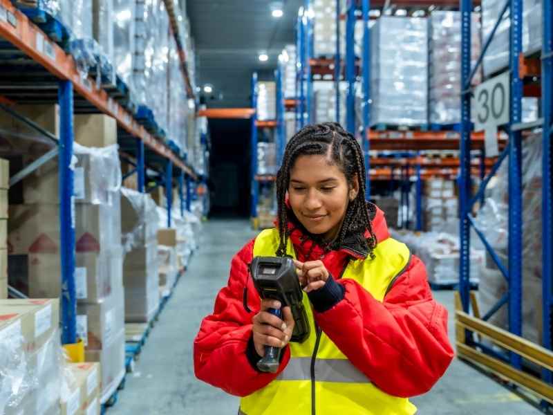 Trabajadora en almacén escaneando productos en estanterías, control de inventario y trazabilidad.