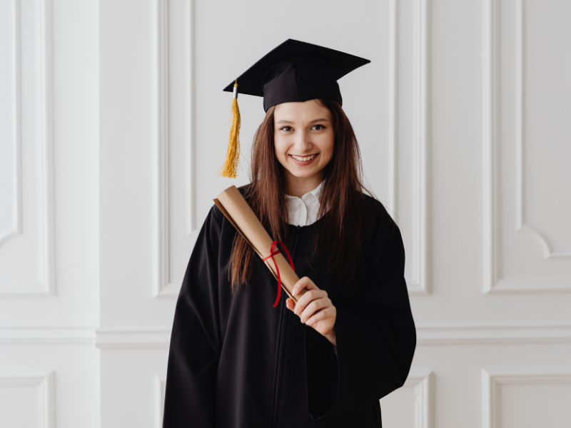 Mujer joven con toga y birrete sonriendo frente a una pizarra con fórmulas, sosteniendo su diploma que representa el éxito de un programa de becas.